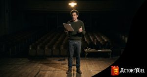 Actor reading from a script under a spotlight on a bare community theater stage during an audition
