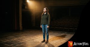 Adult actor standing on a dramatically lit empty stage, taking a deep breath before their first rehearsal